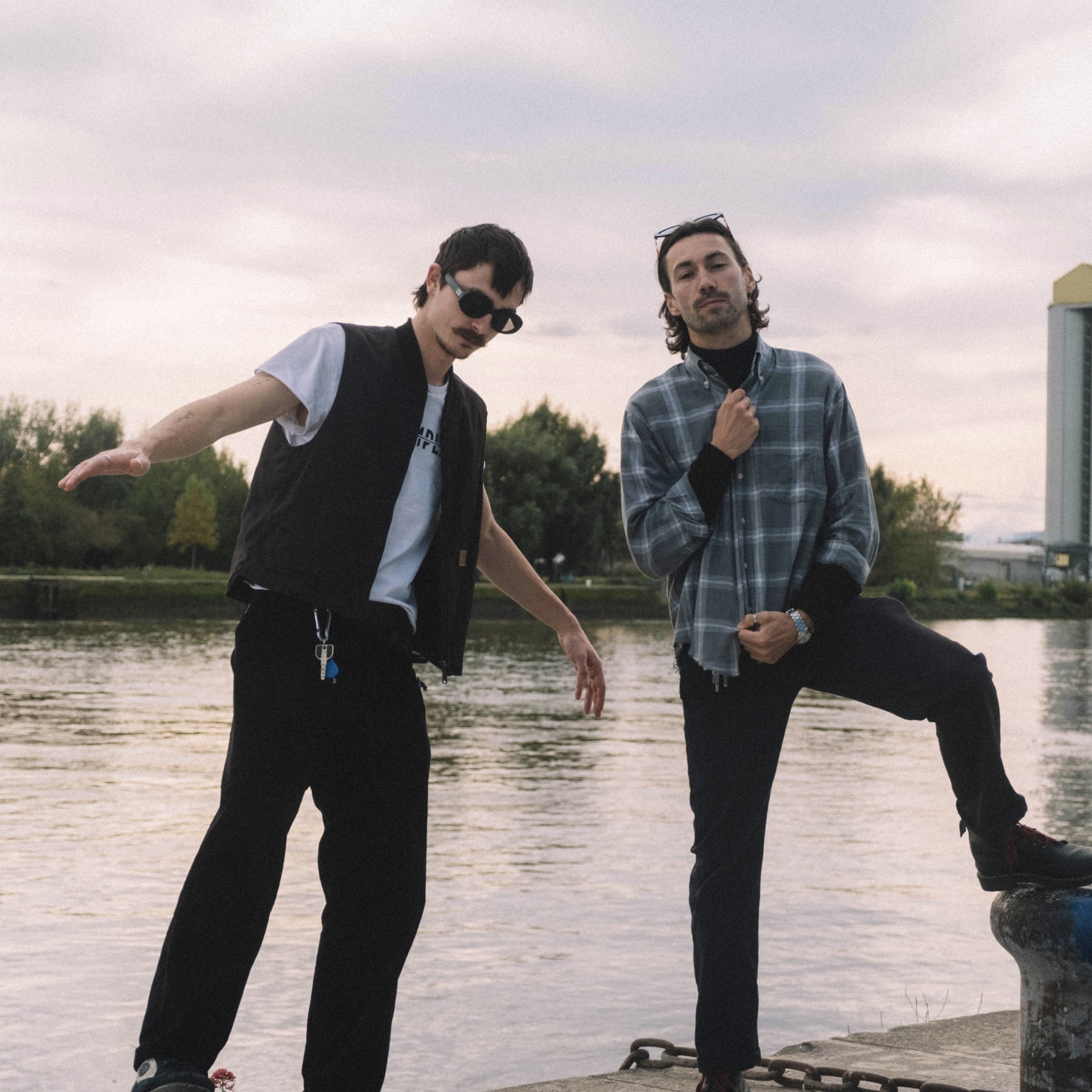 Two men pose by a waterfront at dusk, one in sunglasses and vest, the other in plaid shirt.