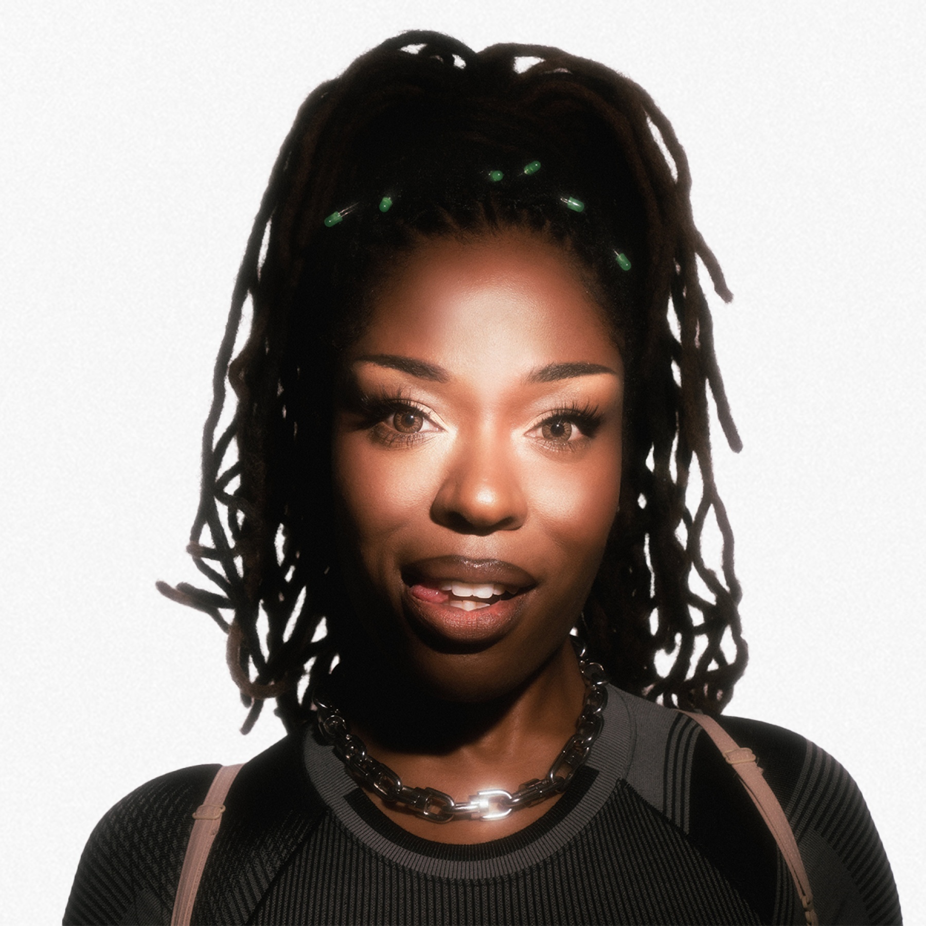 A woman with locs and green accessories wears a black top and chain necklace.