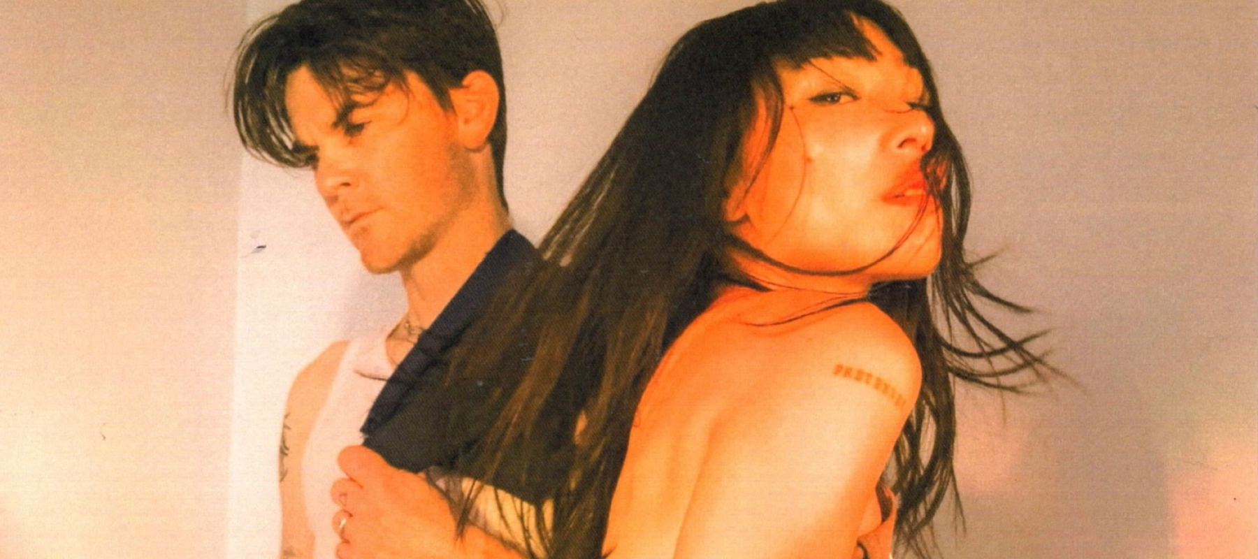 Two people pose back-to-back against a white wall in warm lighting.