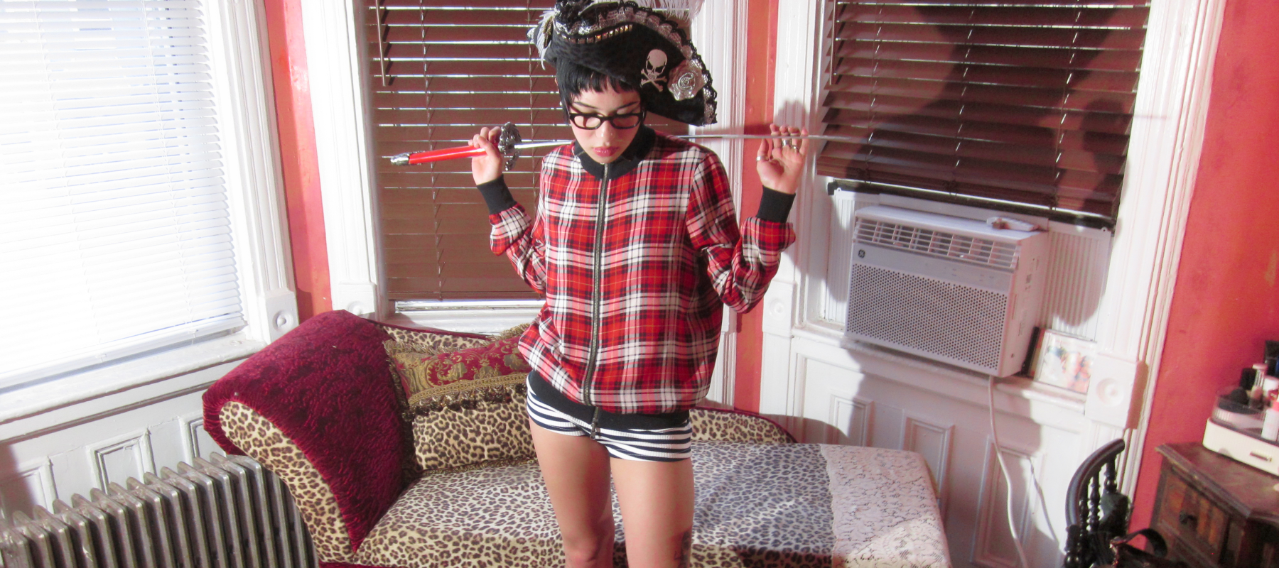 A person in plaid shirt and pirate hat holds a sword in a room with leopard print furniture.