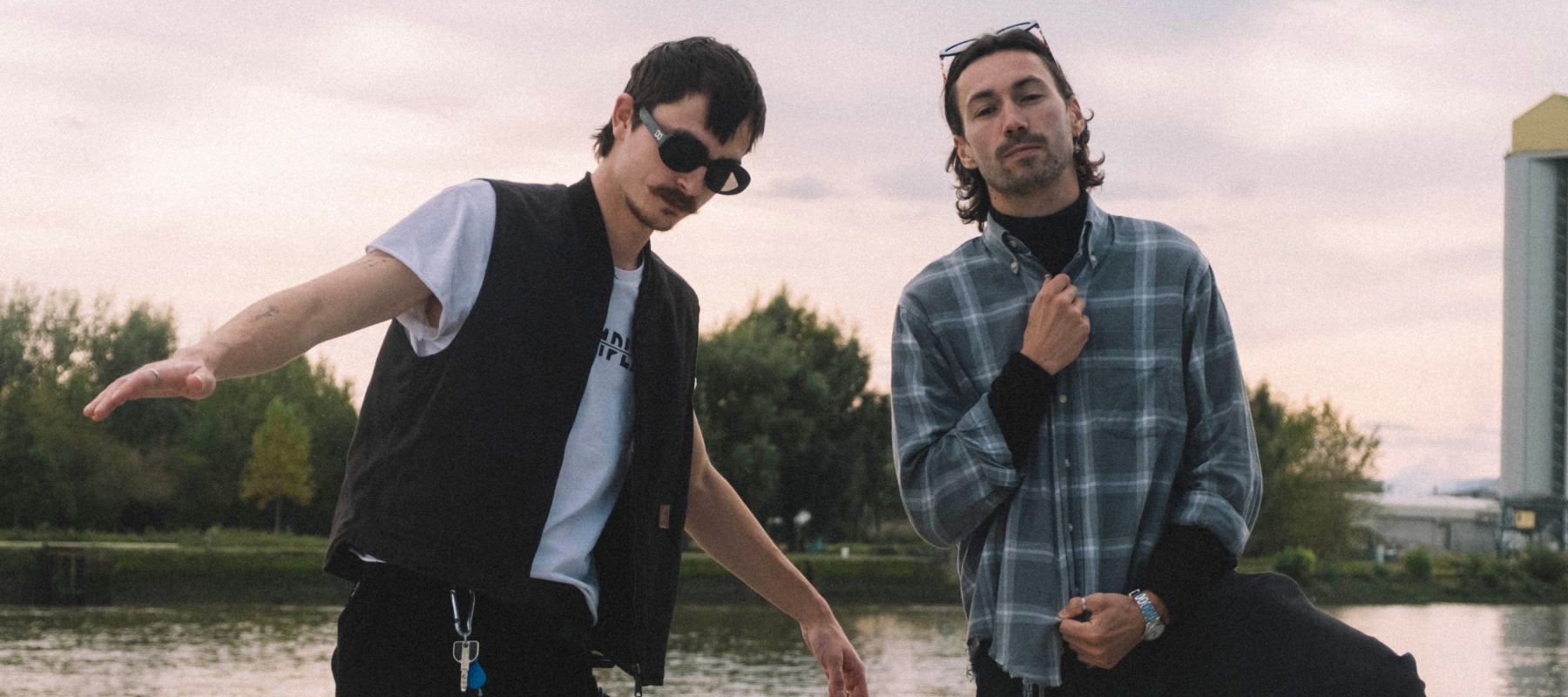 Two men pose by a waterfront at dusk, one in sunglasses and vest, the other in plaid shirt.