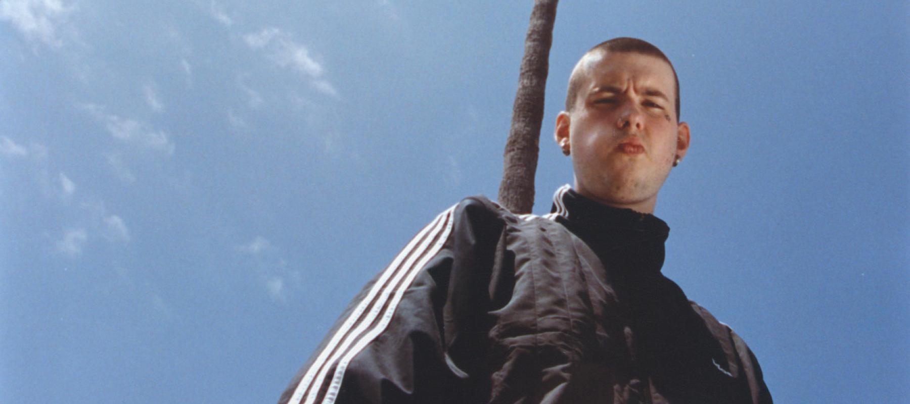 A person in a black tracksuit stands beneath palm trees against a blue sky.