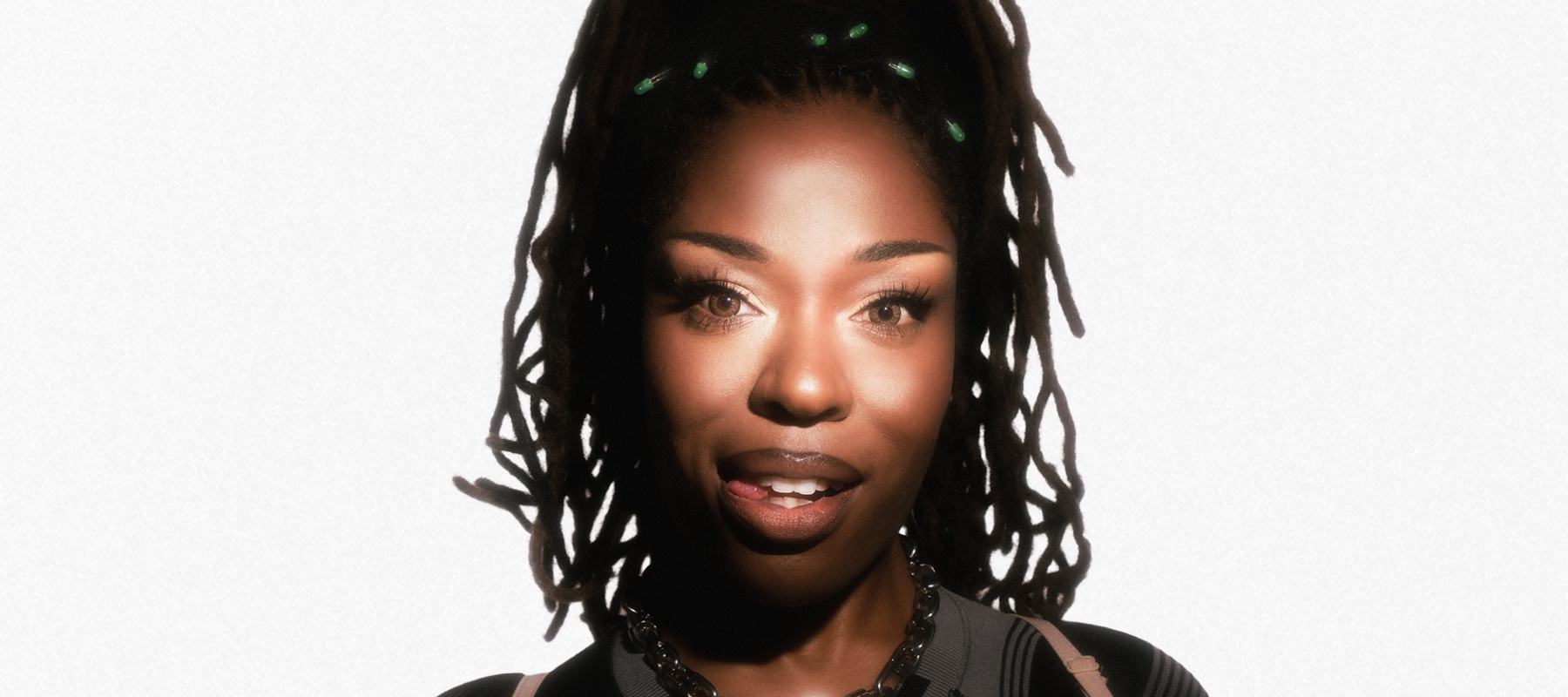 A woman with locs and green accessories wears a black top and chain necklace.