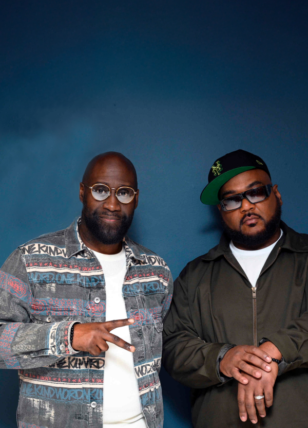 Two men in glasses pose against blue, one in patterned denim, one in black jacket.