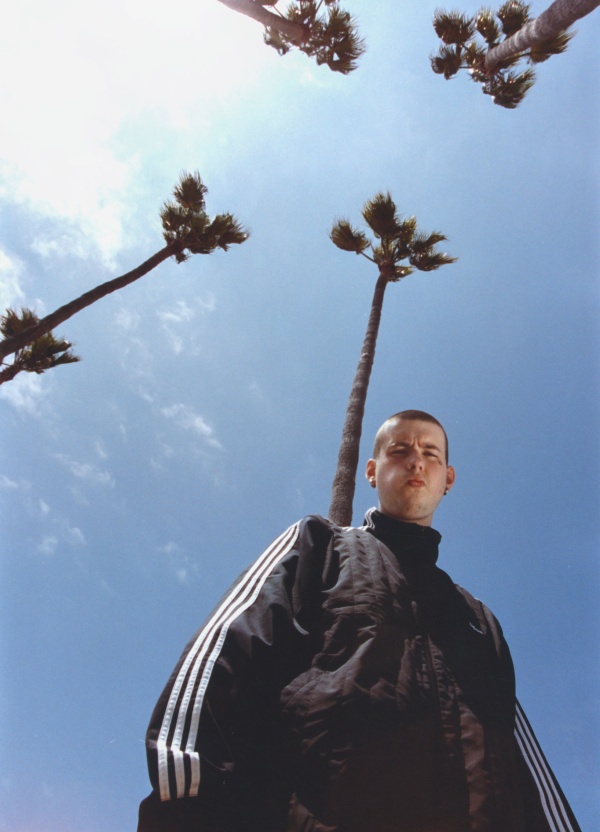 A person in a black tracksuit stands beneath palm trees against a blue sky.