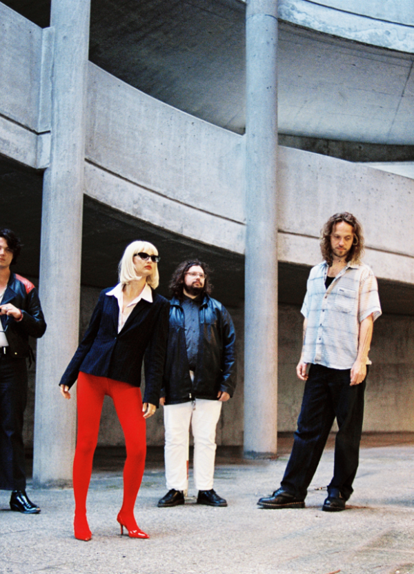 Four people in retro attire pose beneath a brutalist concrete parking structure with angular pillars and ramps.