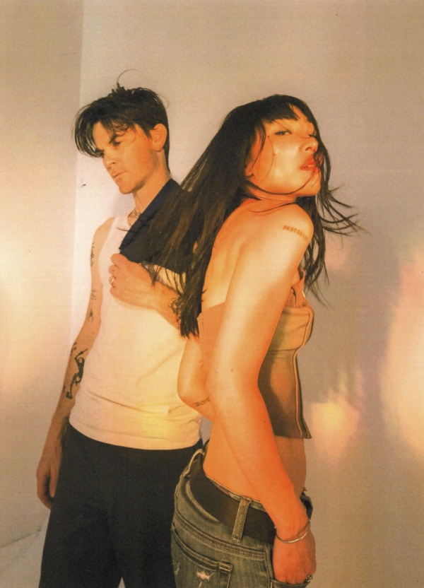 Two people pose back-to-back against a white wall in warm lighting.