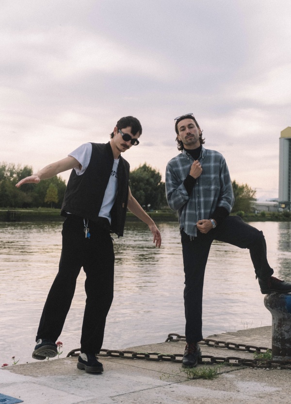 Two men pose by a waterfront at dusk, one in sunglasses and vest, the other in plaid shirt.