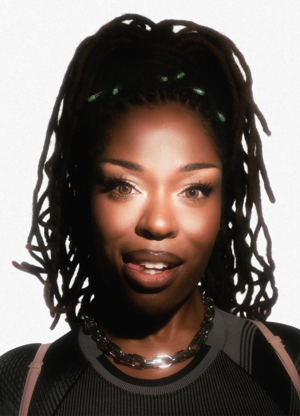 A woman with locs and green accessories wears a black top and chain necklace.